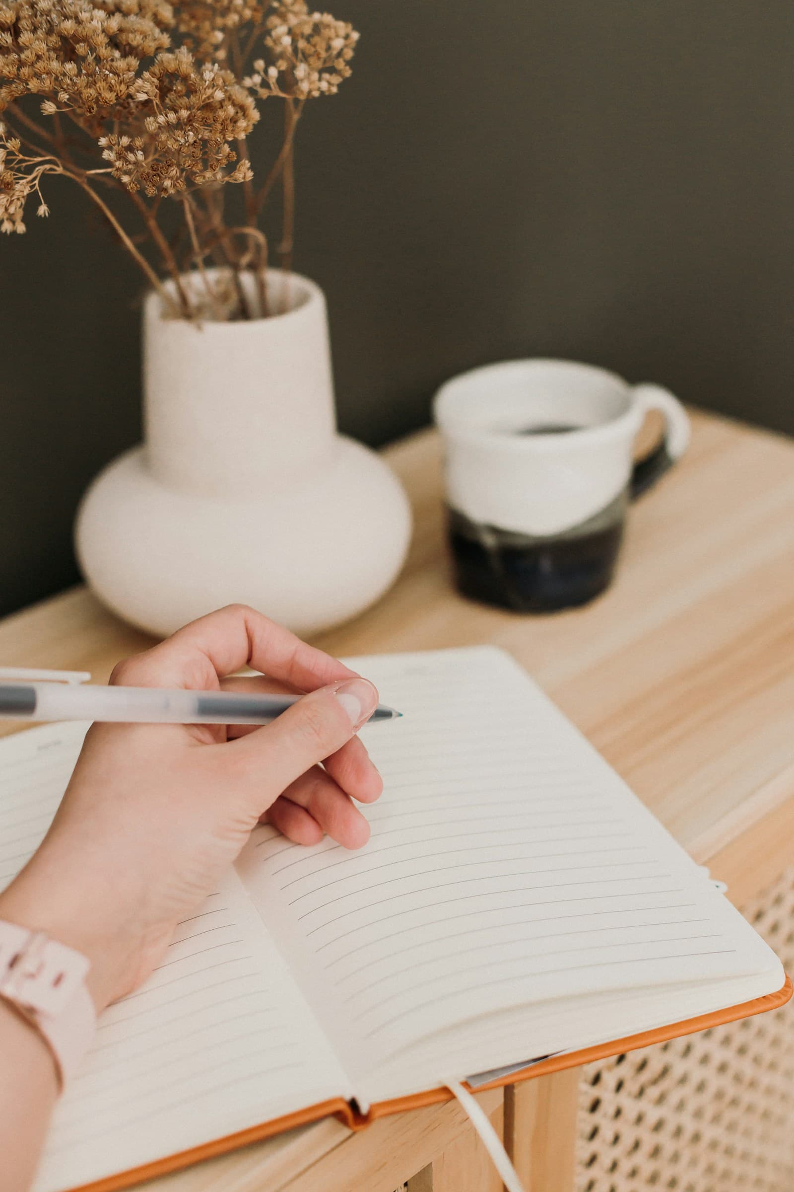Hand writing in a notebook beside a ceramic coffee cup and dried flowers