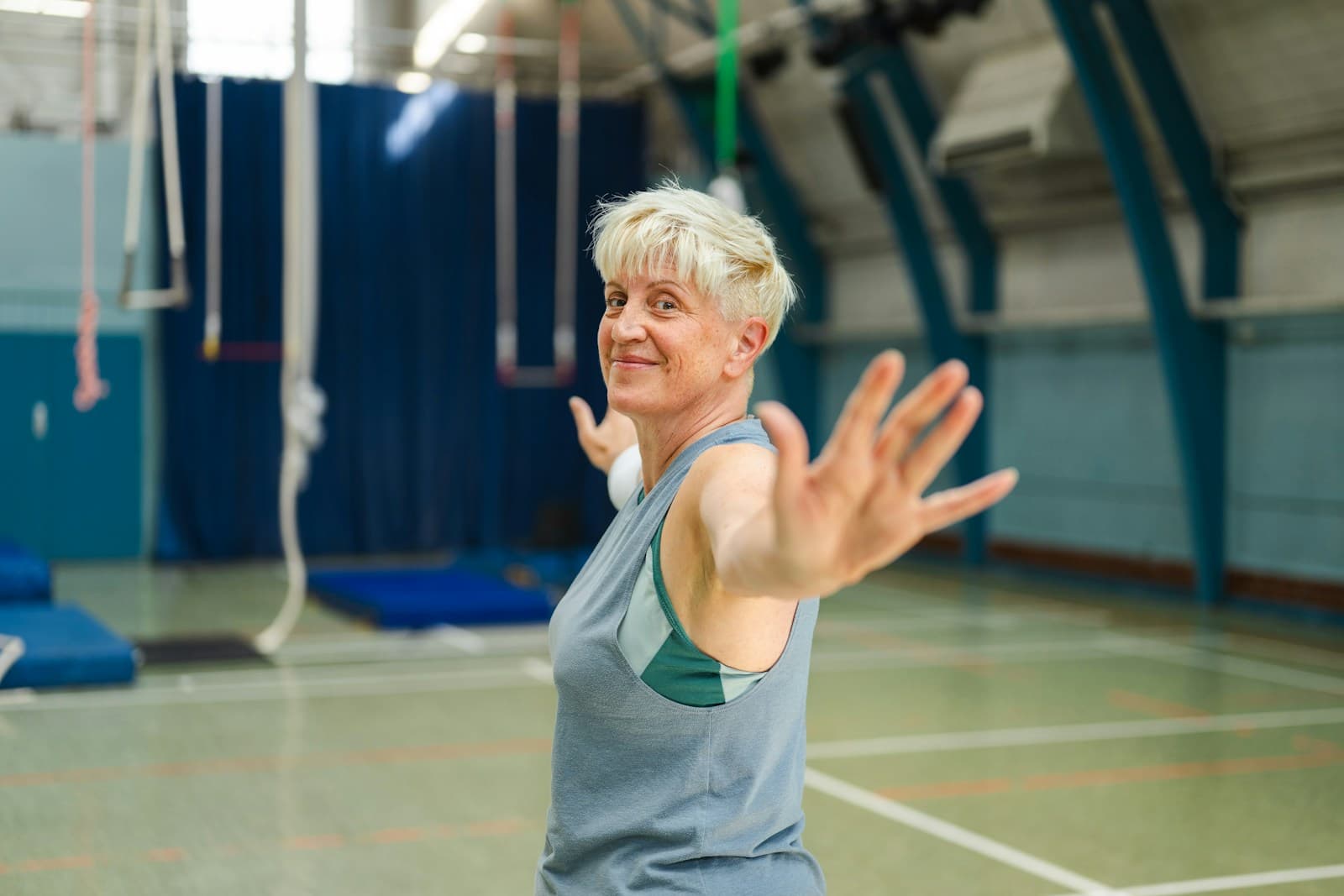An older woman in a gym, mid-stretch, smiling at the camera