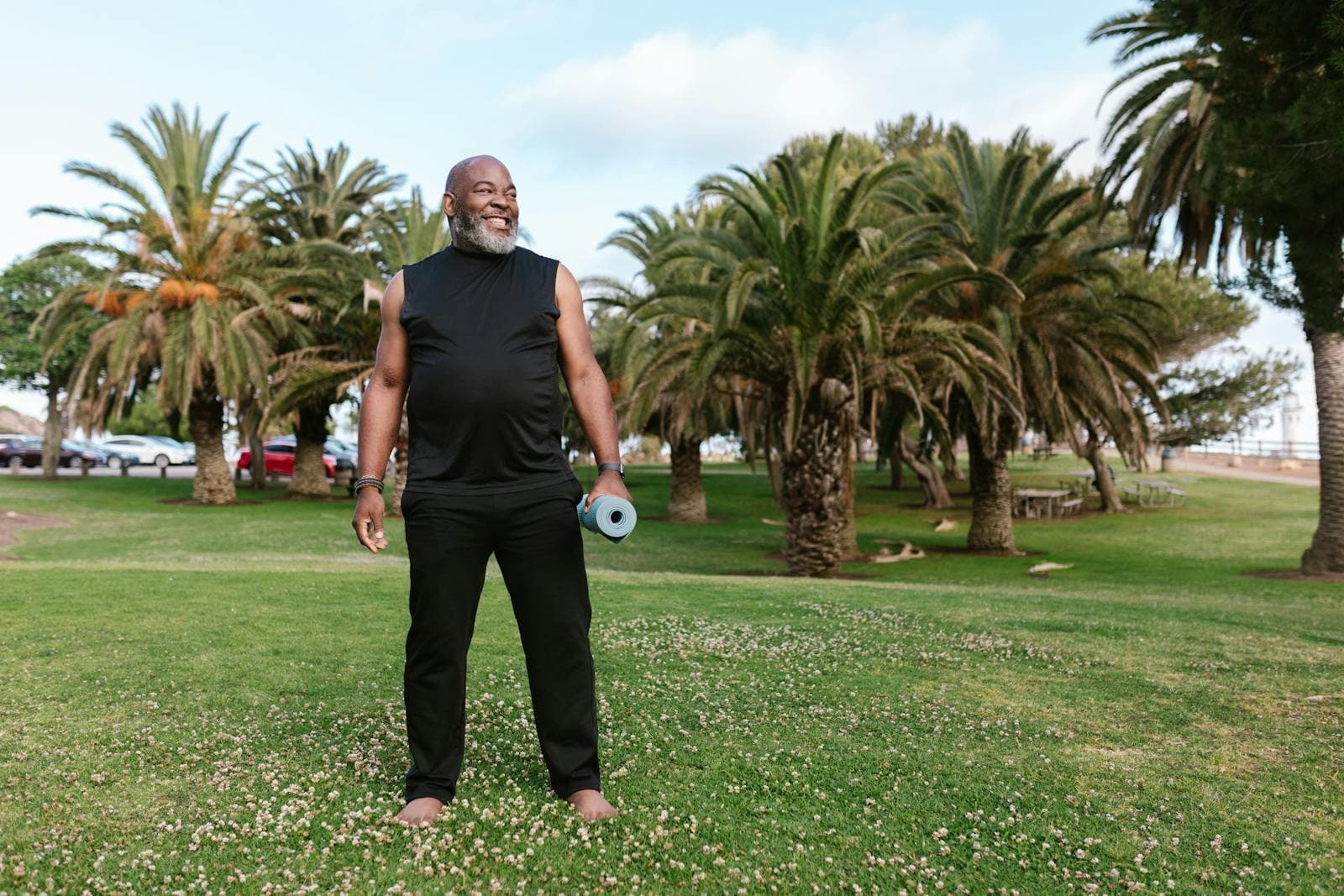An older man smiling in a palm-lined park, holding a rolled yoga mat