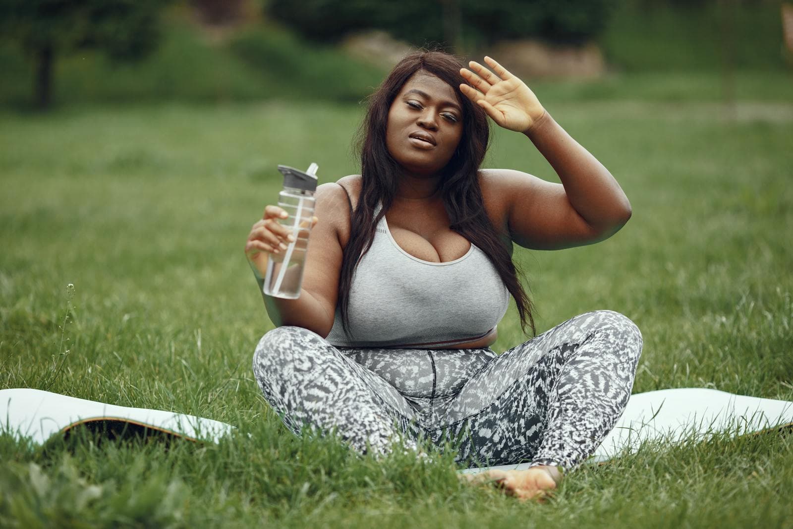 A woman resting on a yoga mat outdoors, wiping her brow and holding a water bottle
